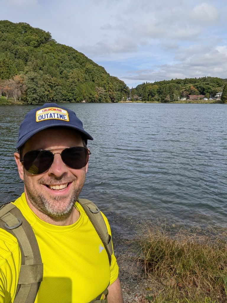Joaby takes a selfie on a lake while on the Shionomichi (Salt Trail) in Japan. He is wearing a bright yellow shirt and a blue hat that says "Outatime" on it.